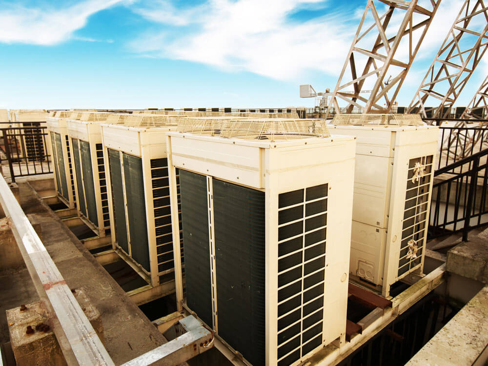Large commercial HVAC condenser units installed on a rooftop, lined up in a row outdoors under a clear sky.