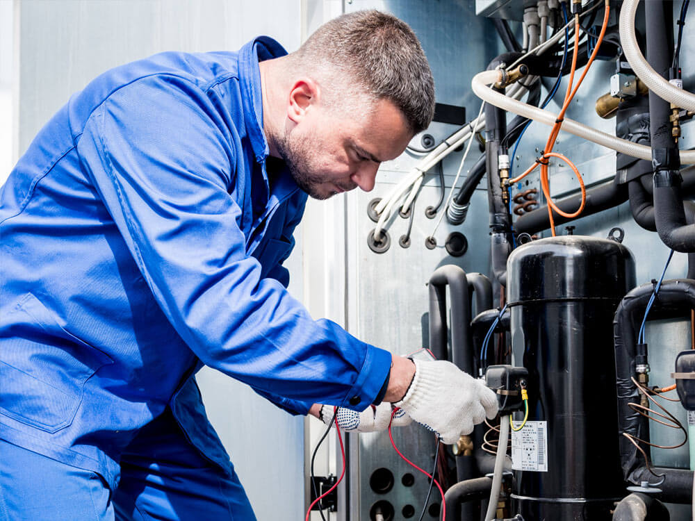 HVAC technician in blue coveralls performs preventative maintenance on a commercial refrigeration compressor, checking wiring and system components.
